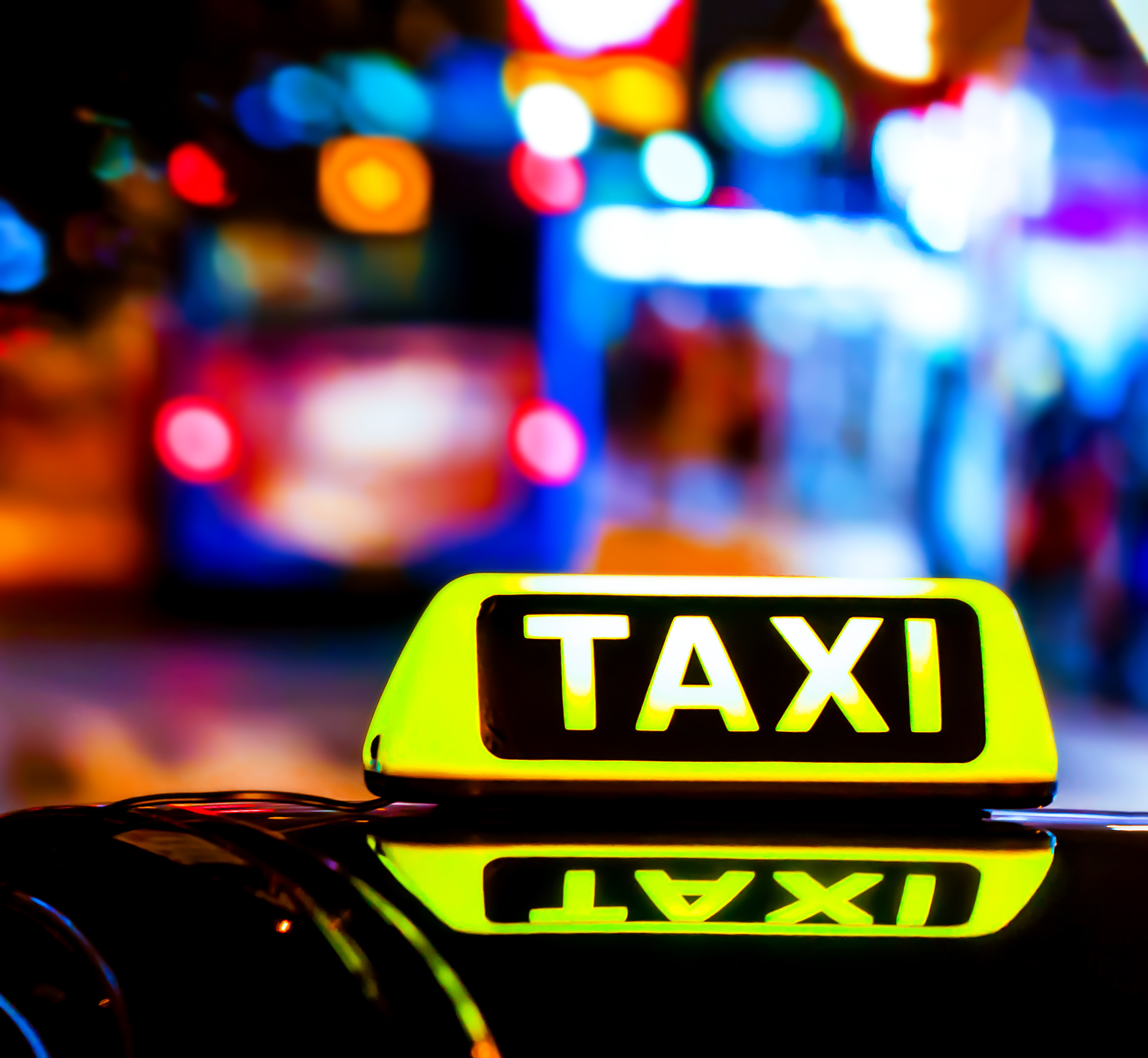 Beleuchtetes Taxi-Schild spiegelt sich auf Fahrzeugdach, nächtliche Straßenszene mit bunten Lichtern unscharf im HIntergrund