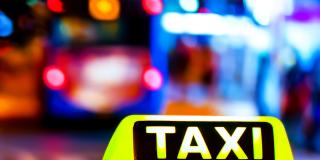 Beleuchtetes Taxi-Schild spiegelt sich auf Fahrzeugdach, nächtliche Straßenszene mit bunten Lichtern unscharf im HIntergrund